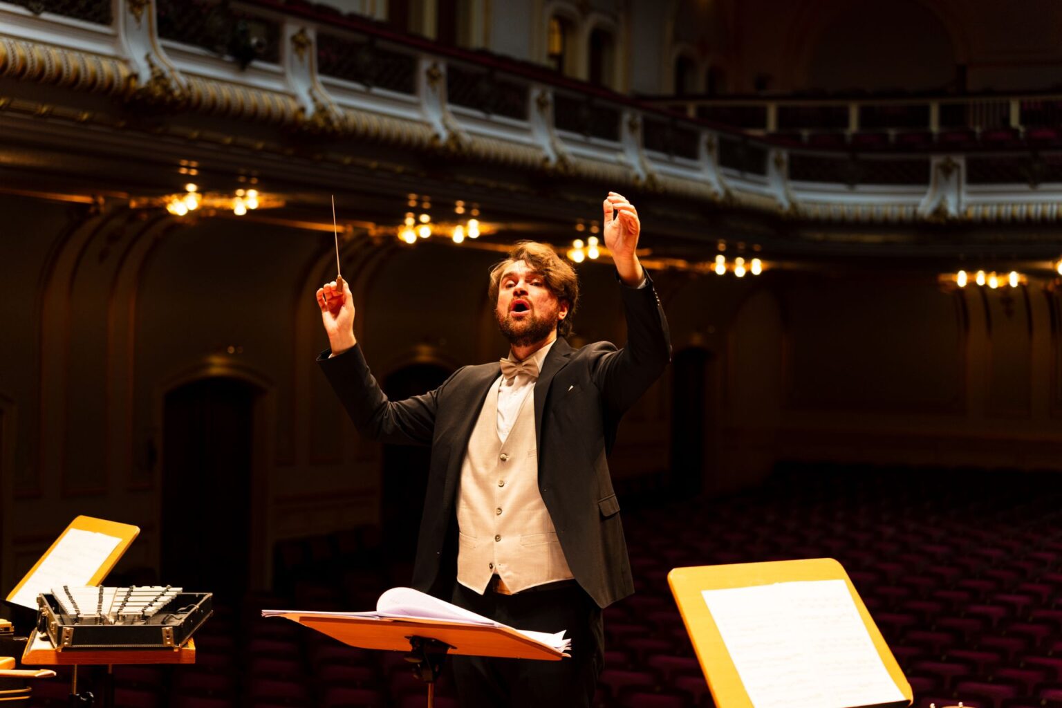 Maestro de casaca rege uma orquestra diante de estantes de partitura em uma elegante sala de concerto iluminada