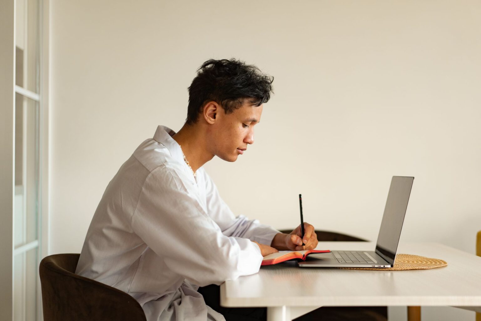Como fazer um TCC sem perder prazos nem arquivos Roupa clara e caderno aberto: jovem sentado à mesa escreve anotações à mão com o notebook aberto ao lado, em ambiente doméstico de estudo