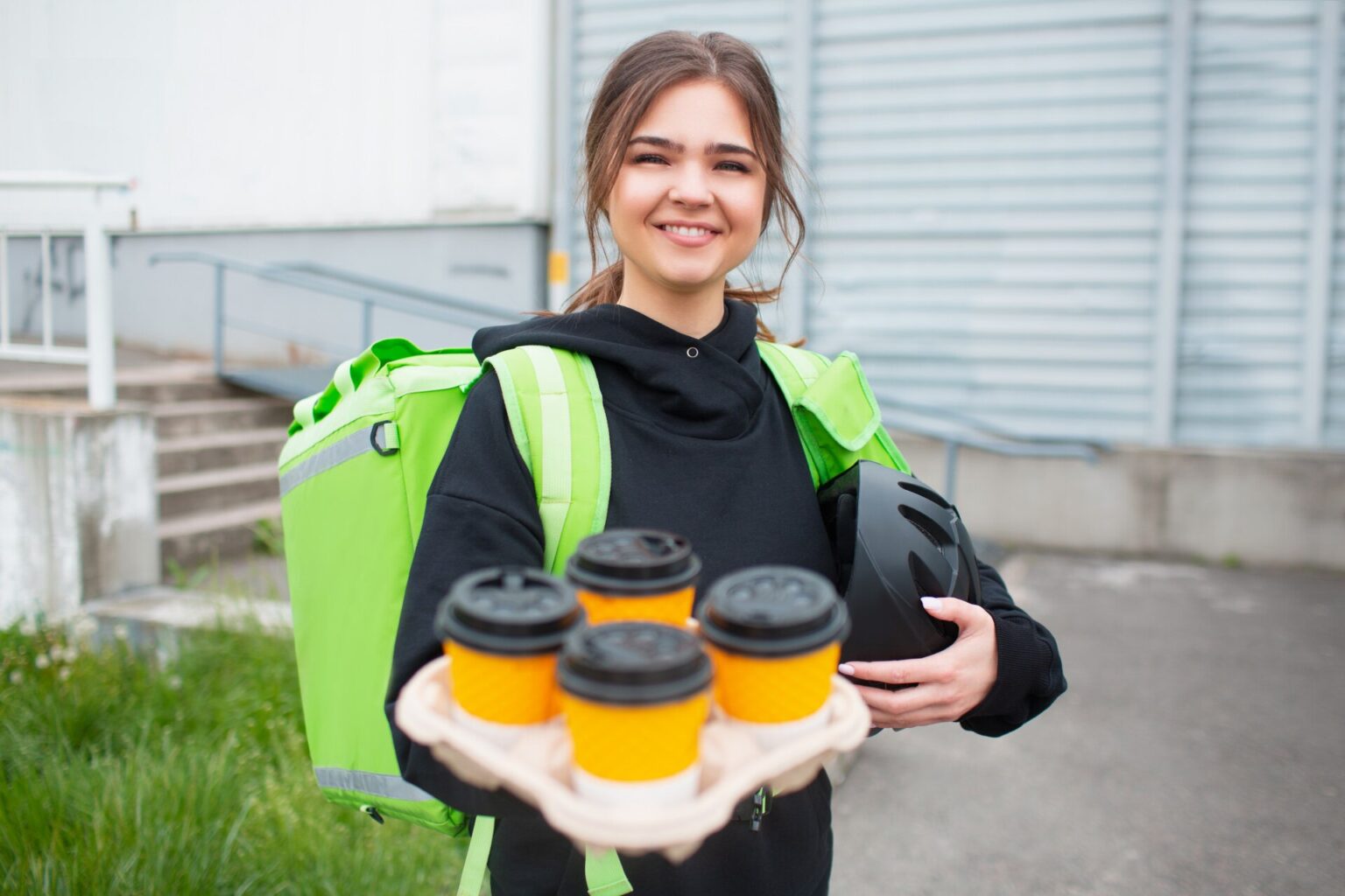 Entregadora sorridente com mochila térmica verde segura bandeja com quatro copos de café tampados e um capacete